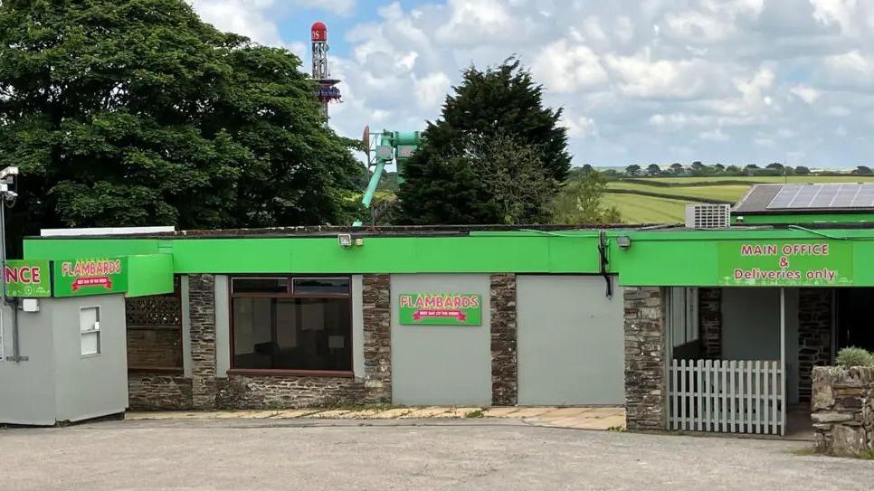 A green and grey building with Flambards branding, some green fields and trees are visible in the background.