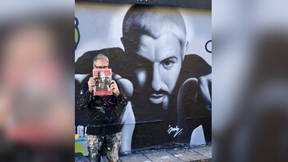 James stands in front of the mural with the front of The Big Issue copied onto the mural. He holds The Big Issue up to his face to cover his nose, with his black rimmed glasses seen over the top of the issue. The front cover of the magazine shows a man pictured in black and white pointing out of the picture.