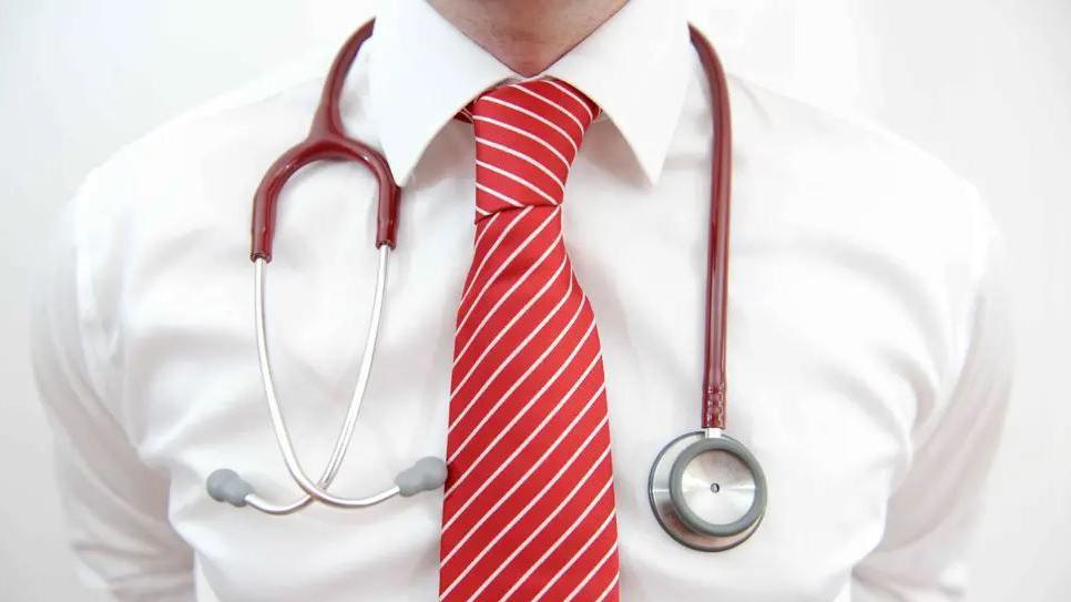 A close up of a man's chest. He is wearing a white shirt and red tie and has a red and silver stethoscope hanging around his neck.