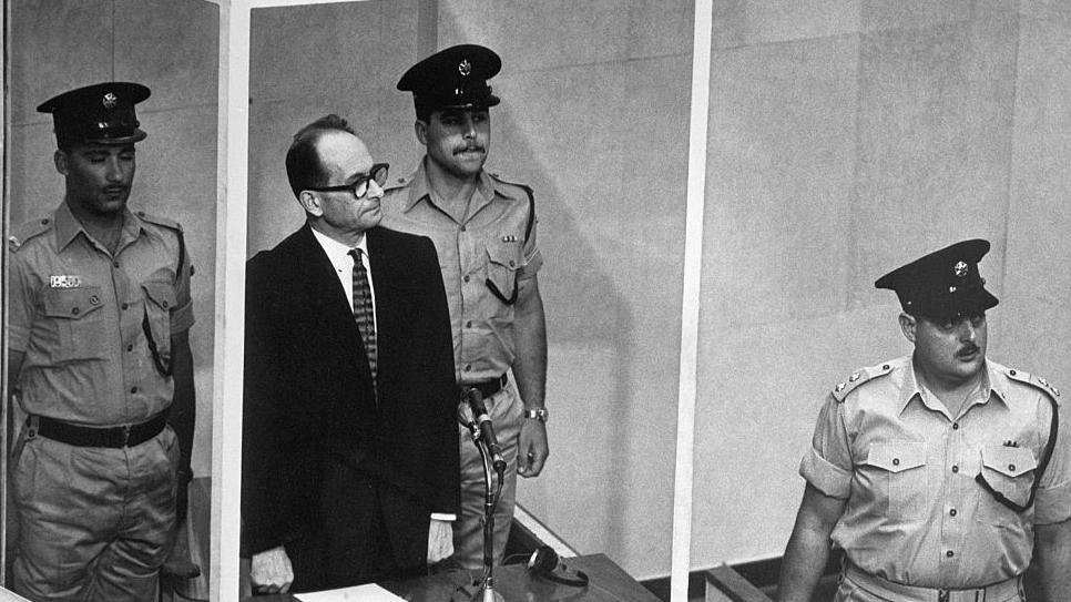 Adolf Eichmann stands in the dock, flanked by two Israeli guards, while a third guard stands at the front right of the picture