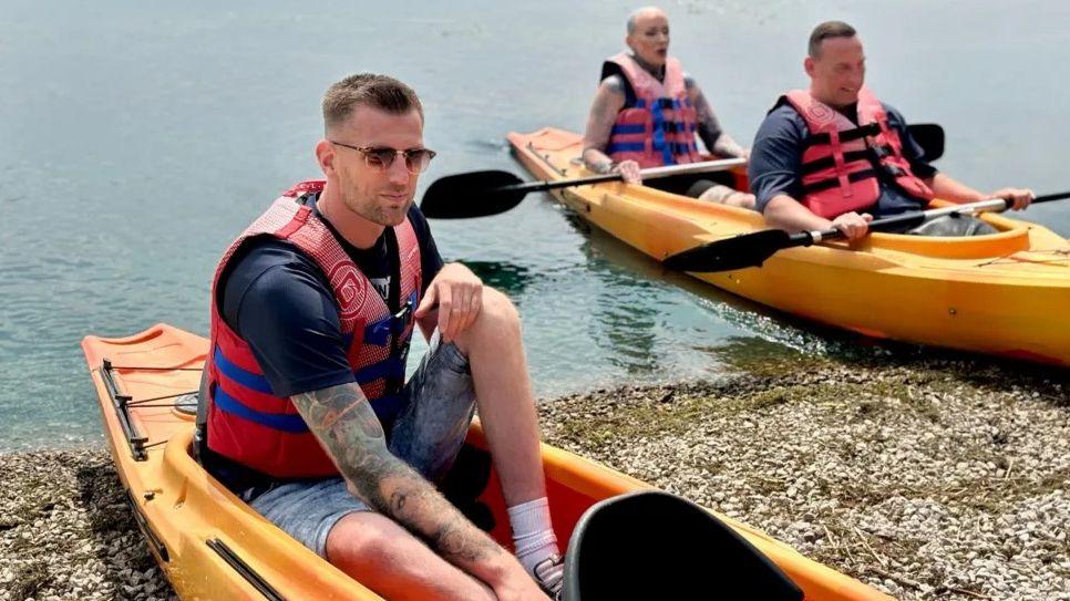 A man in a canoe looks at the camera, with two others in a canoe behind him