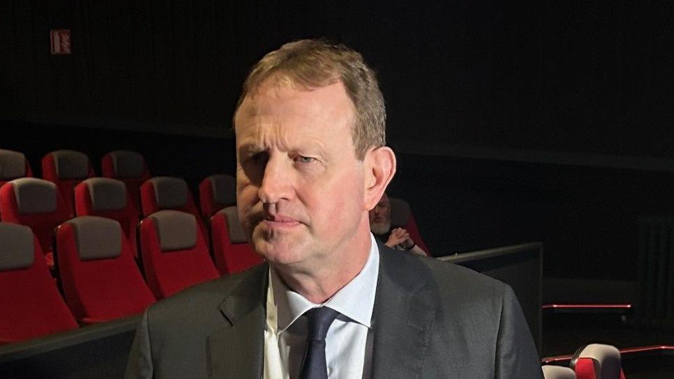 Jim O'Callaghan has fairish/brown short hair and is wearing a dark grey suit jacket, white shirt and dark tie. He is about to respond to a question put to him by a member of the media.