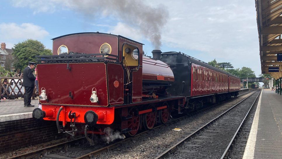 A red and black locomotive and carriages are stood still at a railway station. On the right is a platform and a bridge is in the background.