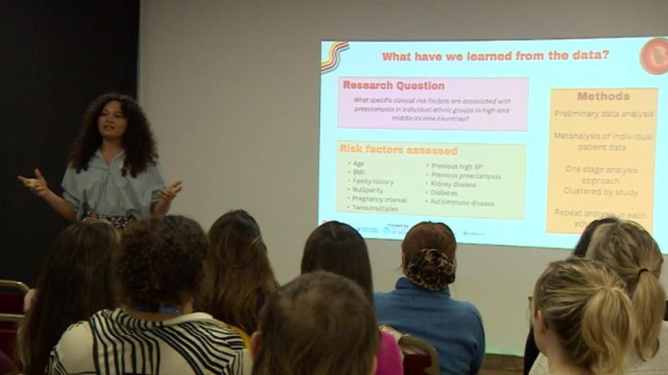 A group of people sit on chairs looking at a projection reading "what have we learned from the data", while a young woman stands at the front leading the talk. 