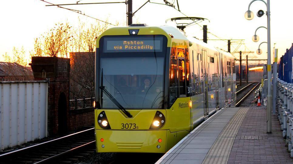 A yellow and grey tram on a track at a platform outside. The sun is setting in the background. 