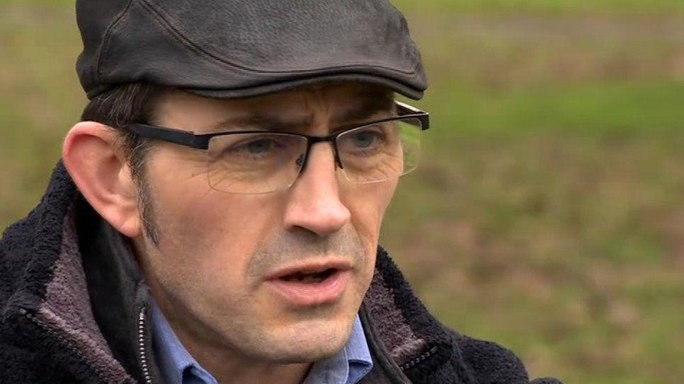 Sean Faloon speaking to the BBC in 2018.  He has short, dark hair and is wearing thin-framed glasses, a black flat cap, a dark fleece and a blue shirt. 