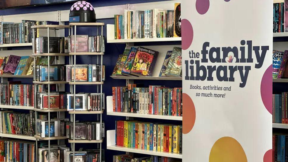 A tall banner reads "The Family Library: books, activities and so much more!" It is white with multicoloured spots and is standing in front of a bookshelf full of books.