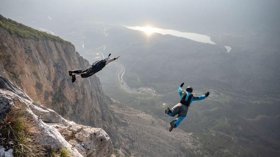 Two people in colourful suits with parachute packs on their backs jump from a cliff. Beyond them is a valley with a green floor and the sun shining off a lake.