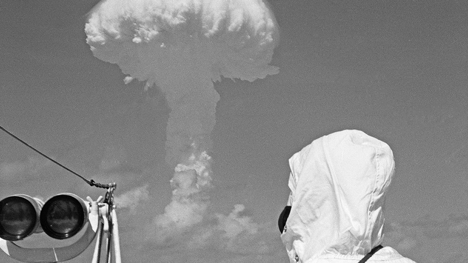 A mushroom cloud rising over the Pacific with a man in protective clothing and goggles in the foreground