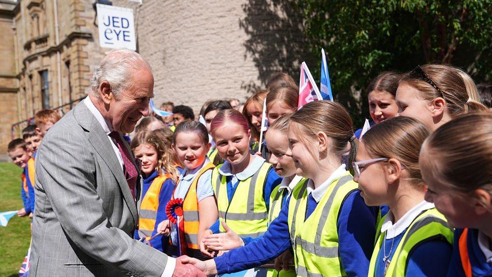 A grey-haired man in a grey suit and red tie shakes hands with a girl wearing a high-visibility vest, while other young girls look on