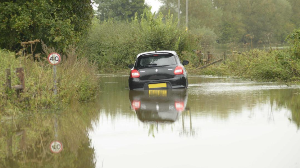 A black car partially submerged, and abandoned on a flooded country road with a partially visible 40 mph sign visible