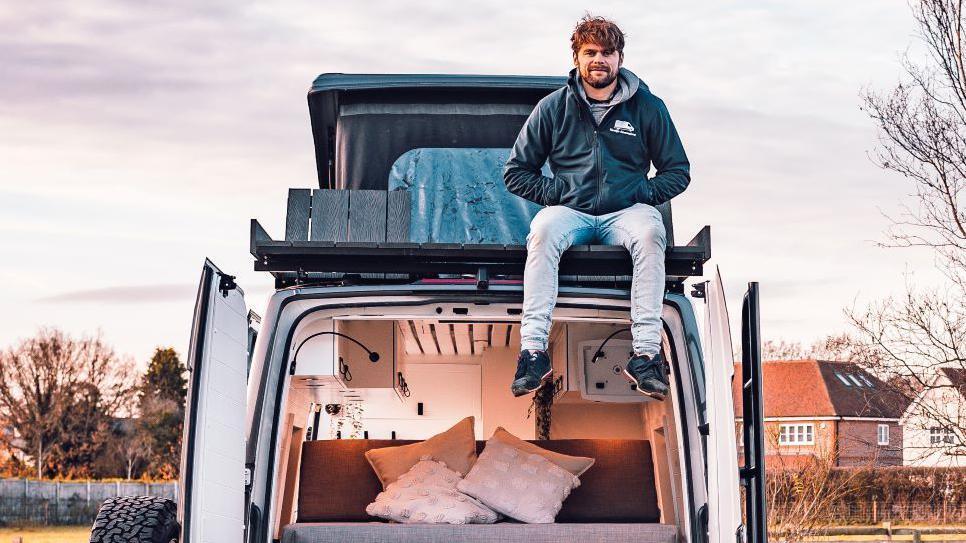 Oli Arnold, who has brown hair and a beard, sitting on top of a van. The doors are open, with a bed and pillows pictured inside.