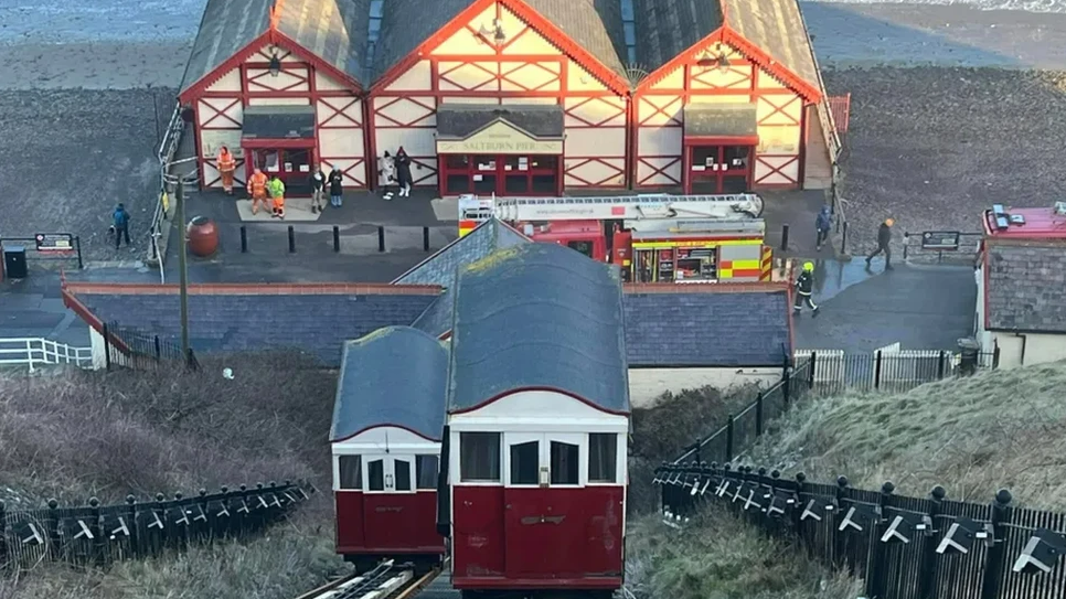 The Saltburn Cliff Lift before it was damaged. At the bottom of the cliff, there is a red and white building with a sign that reads: "Saltburn Pier". In the foreground, two red, white and grey trams can be seen on a rail track ascending/ descending the cliff.