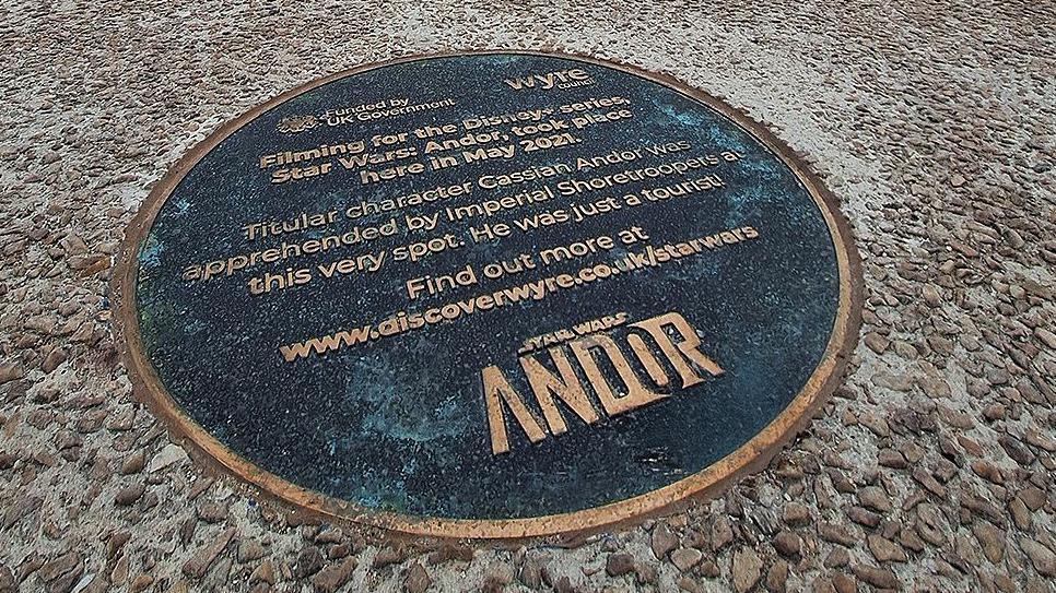 Distance image of the plaque on the promenade showing the cafe in the background and large sandstone wave sculptures to the left