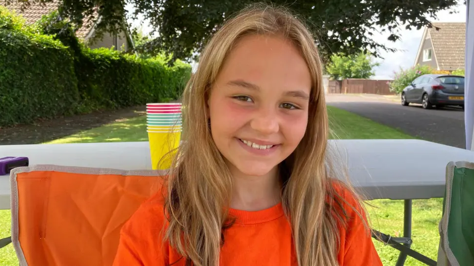 Shyla Brown smiling at the camera as she sits outside on a sunny day wearing a bright orange t-shirt. 