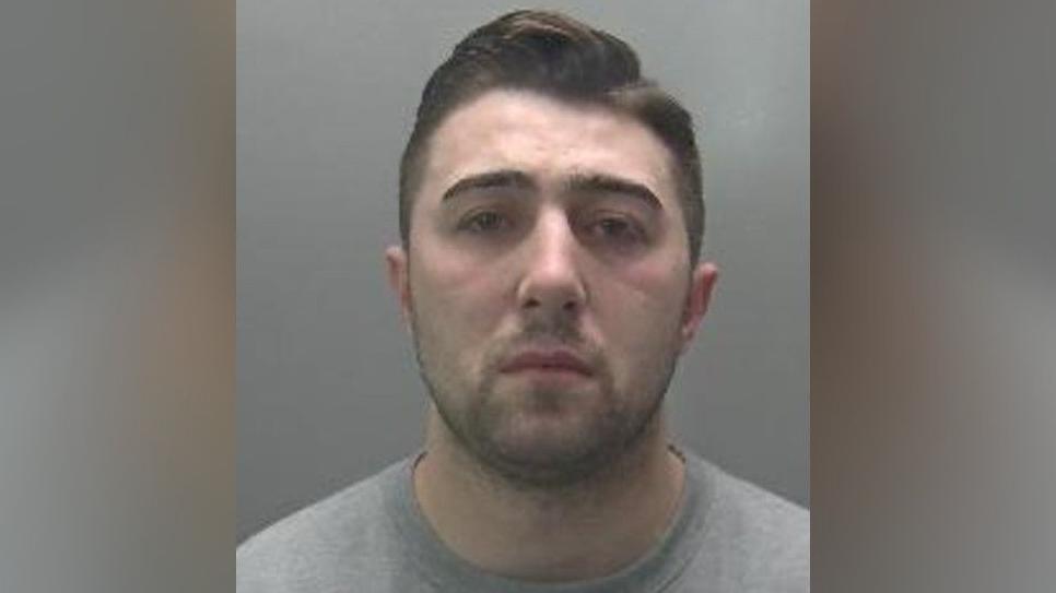 Custody image of Jordan Neale. He has short cropped dark hair and dark stubble, and is wearing a grey jumper.