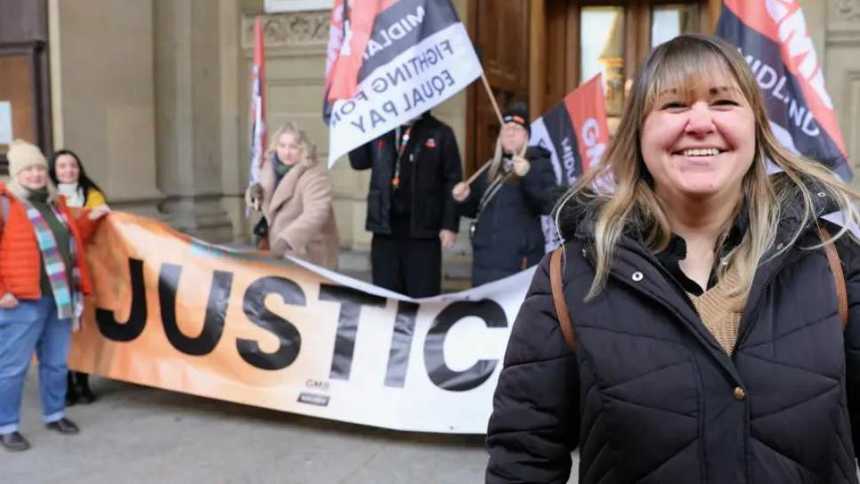 A smiling woman, with shoulder-length hair and wearing a black coat is standing on the right. Behind her are fellow protesters who are holding a banner that says Justice and GMB below it.