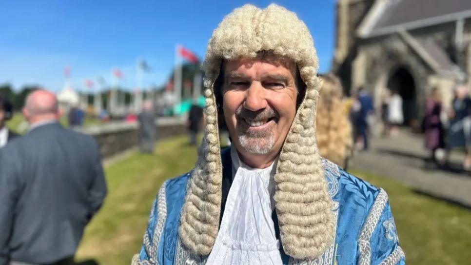 Laurence Skelly smiling ahead of the open-air Tynwald Day ceremony in St John's. He is dressed in the President of Tynwald's blue and white ceremonial robes and wig. A row of flags, other Tynwald Day participants and the Royal Chapel can be seen in the background.