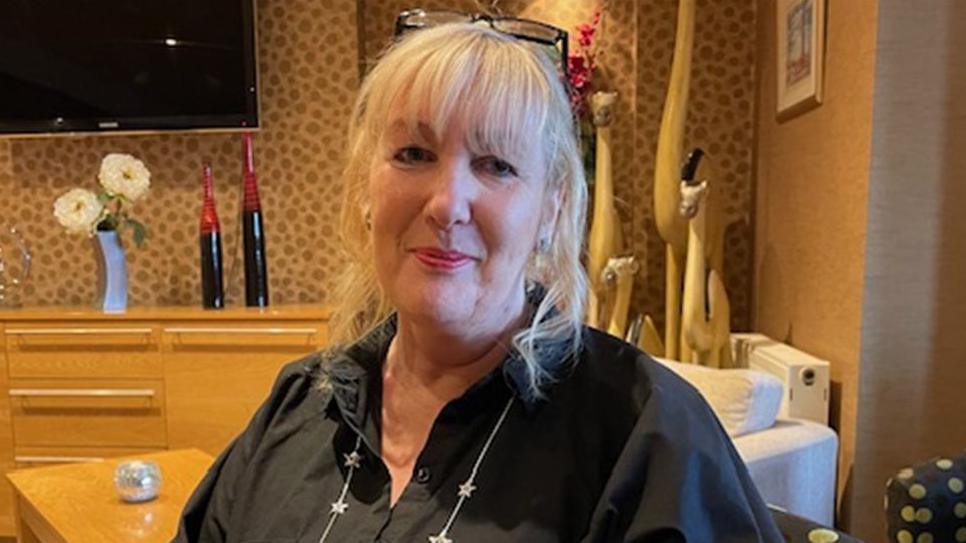 Claire Smith sitting in a hotel room. She is smiling, has long blonde hair and is wearing a black shirt with her glasses on the top of her head