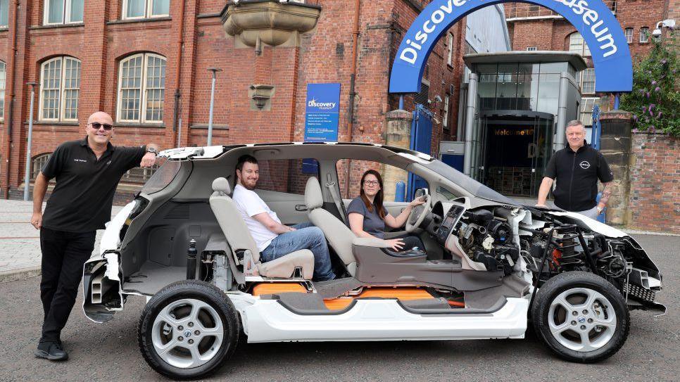 People posed inside and around an electric car outside the Discovery Museum. It is a cross-section so half of the car has been removed, so the interior and inner workings of the engine can be seen.