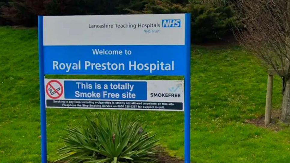 A blue and white sign reading 'Welcome to Royal Preston Hospital' stands amid greenery at the landscaped entrance to the hospital.