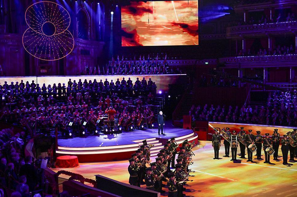 Royal British Legion Festival of Remembrance at the Royal Albert Hall in London. Members of the armed forces are lined up on stage to perform with the outline of a poppy hanging above and rows of audience to each side.