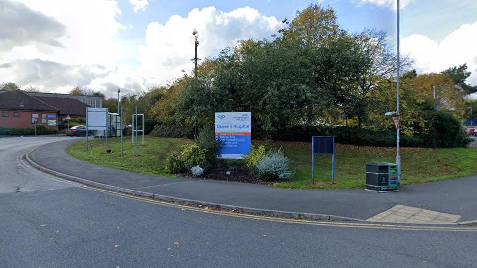 The entrance to Queen's Hospital in Burton. The sign for the hospital is in the centre of the photo, with trees behind it and a path and road in front