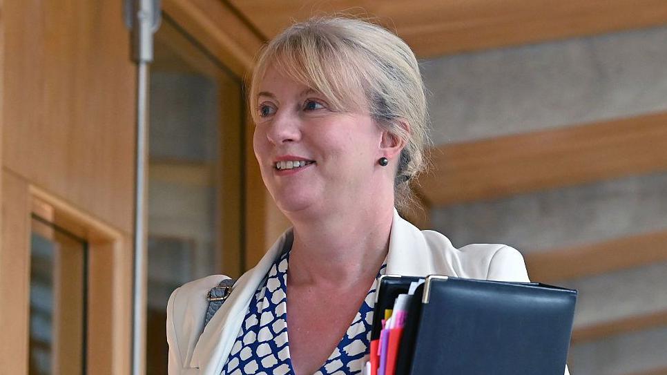 Shona Robison is walking down a wood-panelled corridor in the Scottish Parliament holding a black leather-bound folder. The head-and-shoulders shot shows Robison with her blonde hair tied back and wearing a blue and white top with a white blazer.