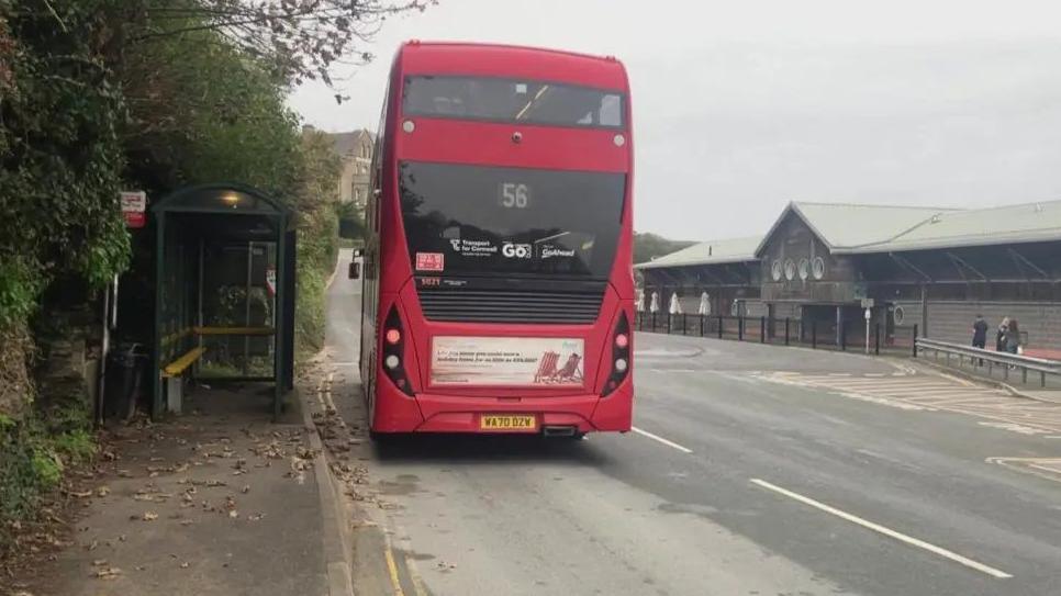 The back of a red double-decker bus, number 56, which is pulled in at the bus stop on Station Road. A wooden building is opposite the bus stop across the road. 