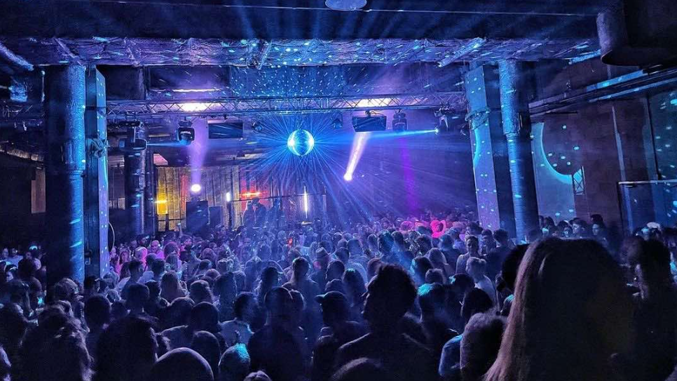 Hundreds of people enjoy a dance event at The Underground in Bristol. They are illuminated by blue and white lights and light beams are shooting out from a glittering disco ball