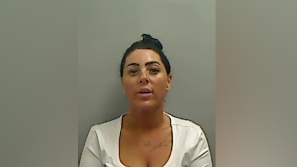 Mugshot of Simone Reed. The image is pixilated but it can be seen that she has black hair scraped into a bun, thick black eyebrows and false eyelashes. She has tattoos across her collar bone and is wearing a low cut white top.