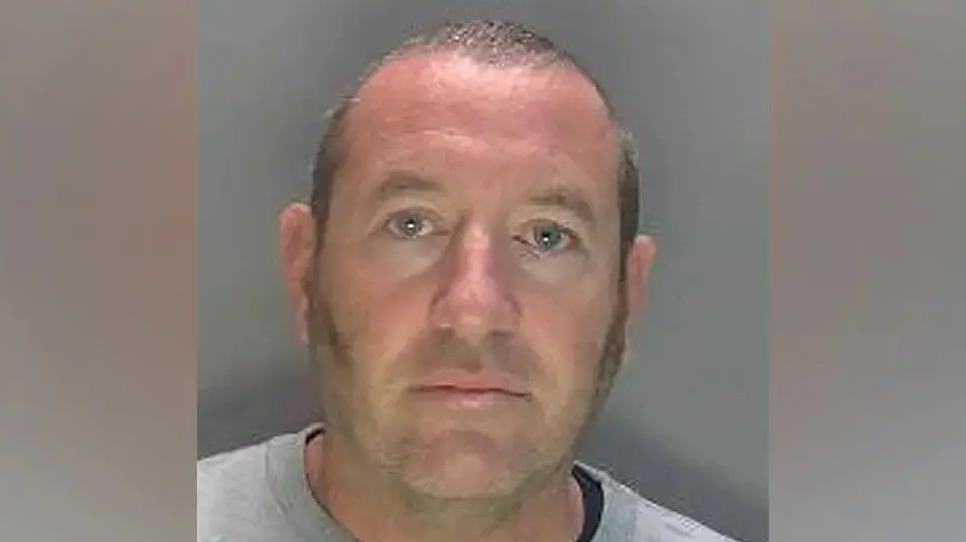 David Carrick photographed in police custody. He is wearing a grey sweatshirt and has sideburns and a near-shaven head. 