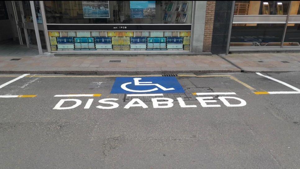 A parking space on a road with a logo of a person in a wheelchair and the word DISABLED. Above the pavement are two glass-fronted shops.
