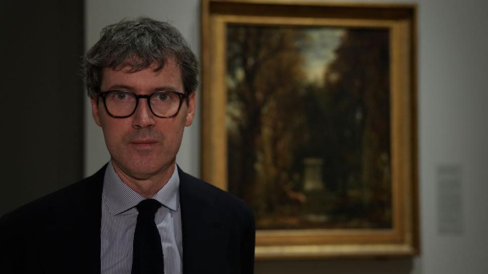 A man, Per Rumberg, is standing in front of a painting inside a gallery. He is wearing a striped shirt, black jacket and tie and glasses.