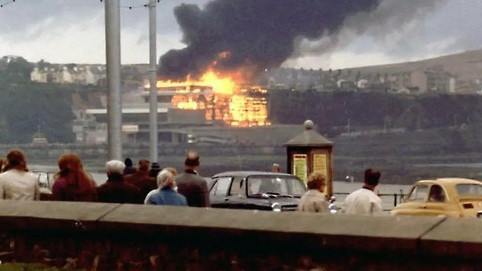 A group of people look on as the fire takes hold of the Summerland complex with black smokie rising above it.