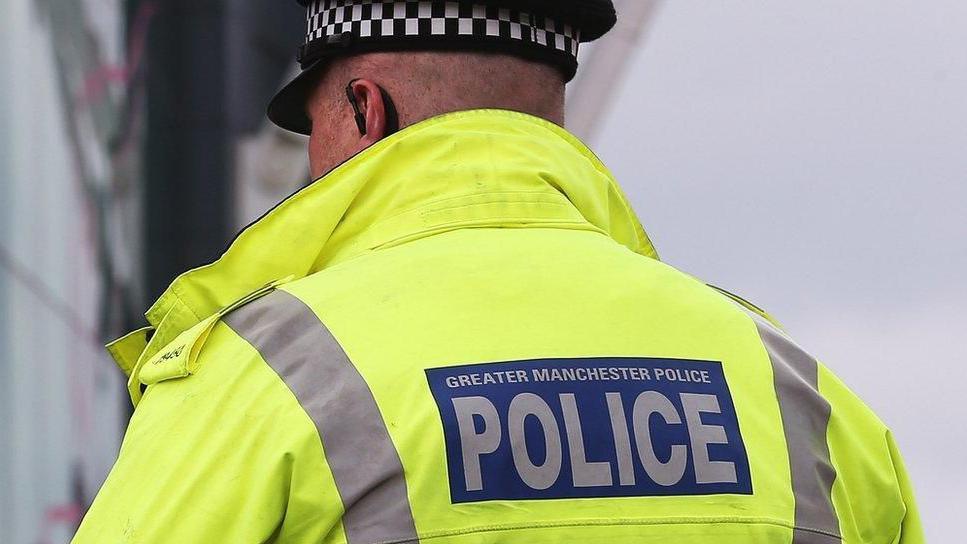 The image shows the back of a police officer wearing a bright fluorescent yellow jacket. The jacket has a large rectangular blue patch on the back with white text that reads “GREATER MANCHESTER POLICE”, with the word “POLICE” in bold, capital letters. The officer is also wearing a black-and-white checkered hat, which is partially visible at the top of the image. The background is blurred, but it appears to be outdoors in daylight, with muted colors suggesting a cloudy sky.