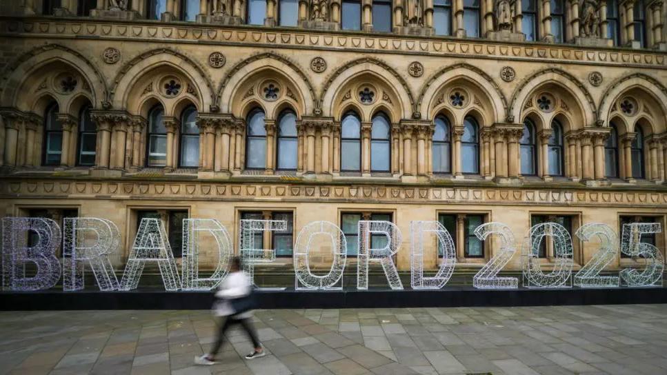 A person walks past Bradford City Hall with a Bradford 2025 sign in front of it. The sign has large white 3D letters and is sitting on a dark base. 