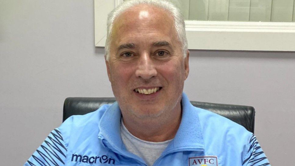 A man with white hair with a light blue top containing the letters AVFC for Aston Villa football club. He is sitting on a black chair with a wall in the background.