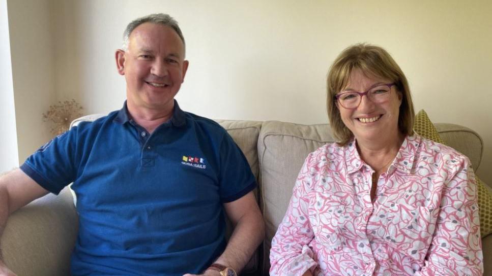 David Lowe with short grey hair in a blue polo shirt and shorts, and Hilary Lowe in a floral pink and white shirt and pink glasses, sitting on the sofa and smiling at the camera