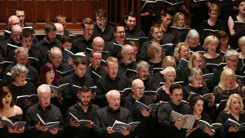 Shows a choir of men and women dressed in black singing 