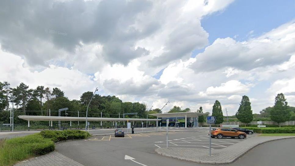 Station car park, with three parked cars.  There are walkways covered by white roofs and trees in the background. There is a cloudy sky overhead. 