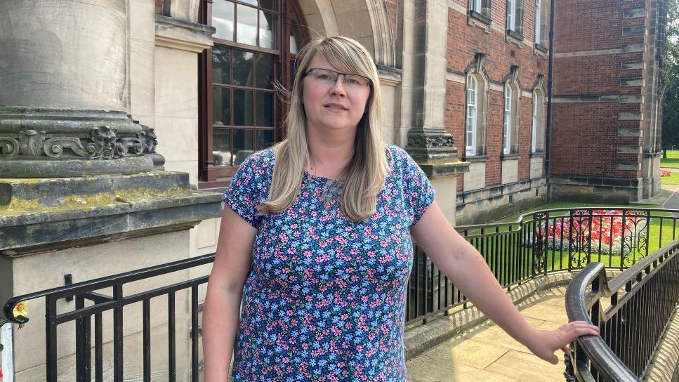 Picture shows a woman with long mousy hair, wearing a flowery short sleeved top and glasses stood in front of a town hall with her left hand resting on the black railings