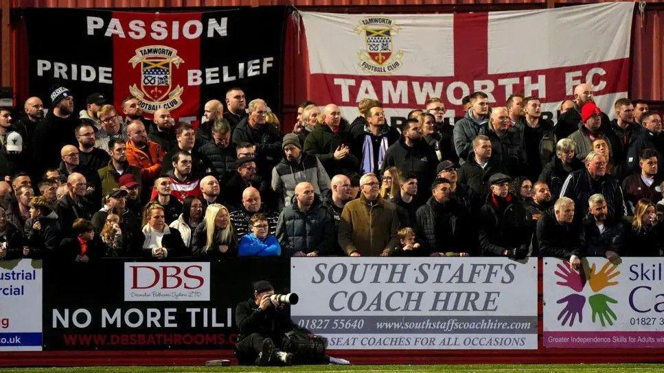 Tamworth fans in the stands. There are sponsorship boards around them and flags in the background.