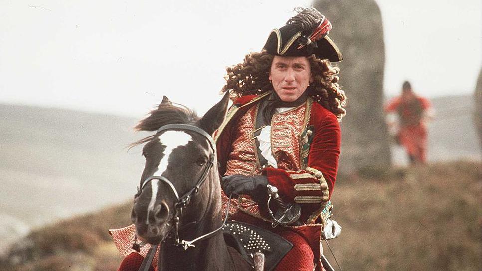 Tim Roth in powdered wig and red coat rides a horse in Rob Roy, an action shot with a hill behind him and other soldiers on the grass.