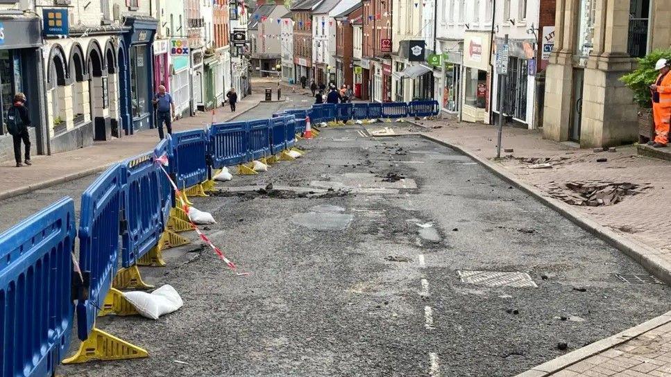 A street with damaged road and pavement is cordoned off by plastic barriers. The street is lined with rows of shops.