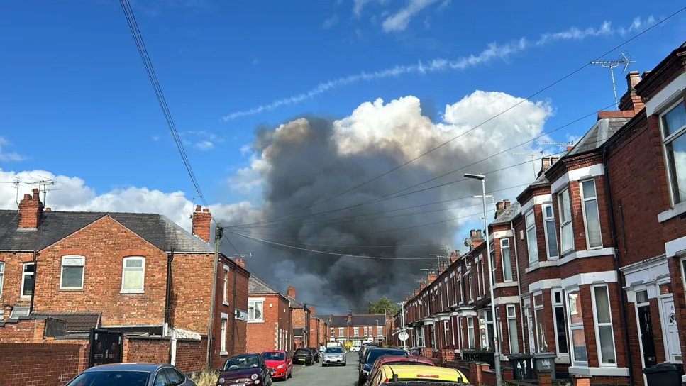 Plumes of dark grey smoke rise from behind a street of red-brick houses