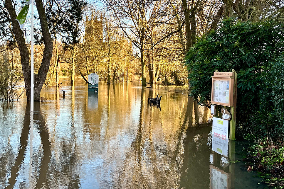 Flooded Worcester. Paths are covered with water, with the benches and signs part-submerged. The Cathedral is in the distance