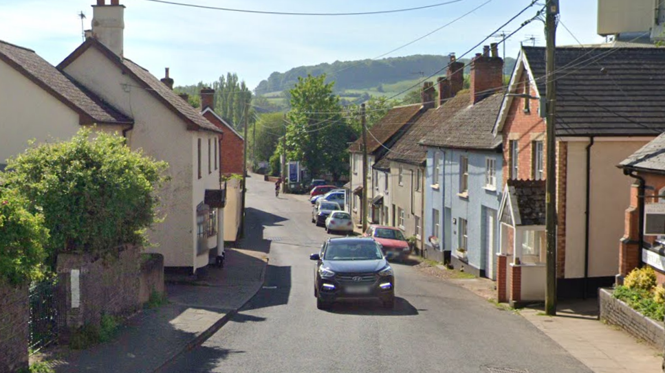 Google image of Bridge Street in Uffculme. Residential homes are on either side of the narrow street. Cars are parked on the right side of the road. A car is travelling up the road.