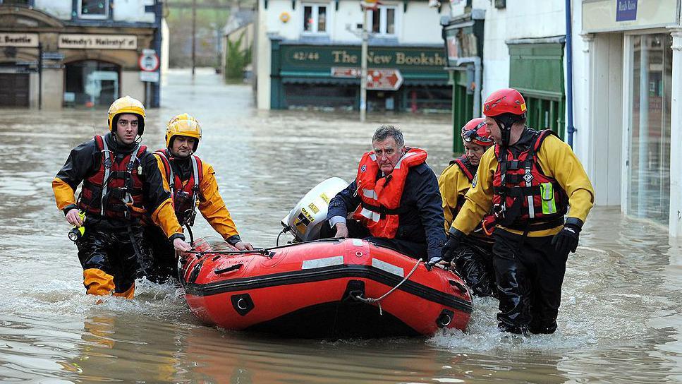 Emergency rescue workers use an inflatable boat to rescue a stranded man in Cockermouth town centre. The flood waters are several feet deep. The four crew members are wearing protective clothing while the man in the boat has a life vest.
