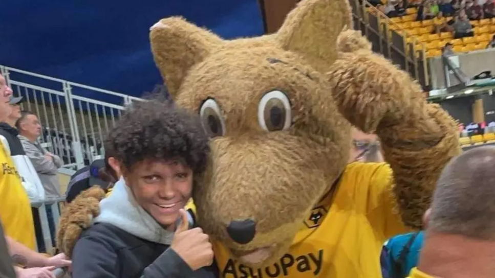 Harleigh is smiling with his thumb stuck up and arm round a football mascot at a Wolves match. He has long, curly black hair. The Wolves mascot is a big furry wolf wearing a gold top.